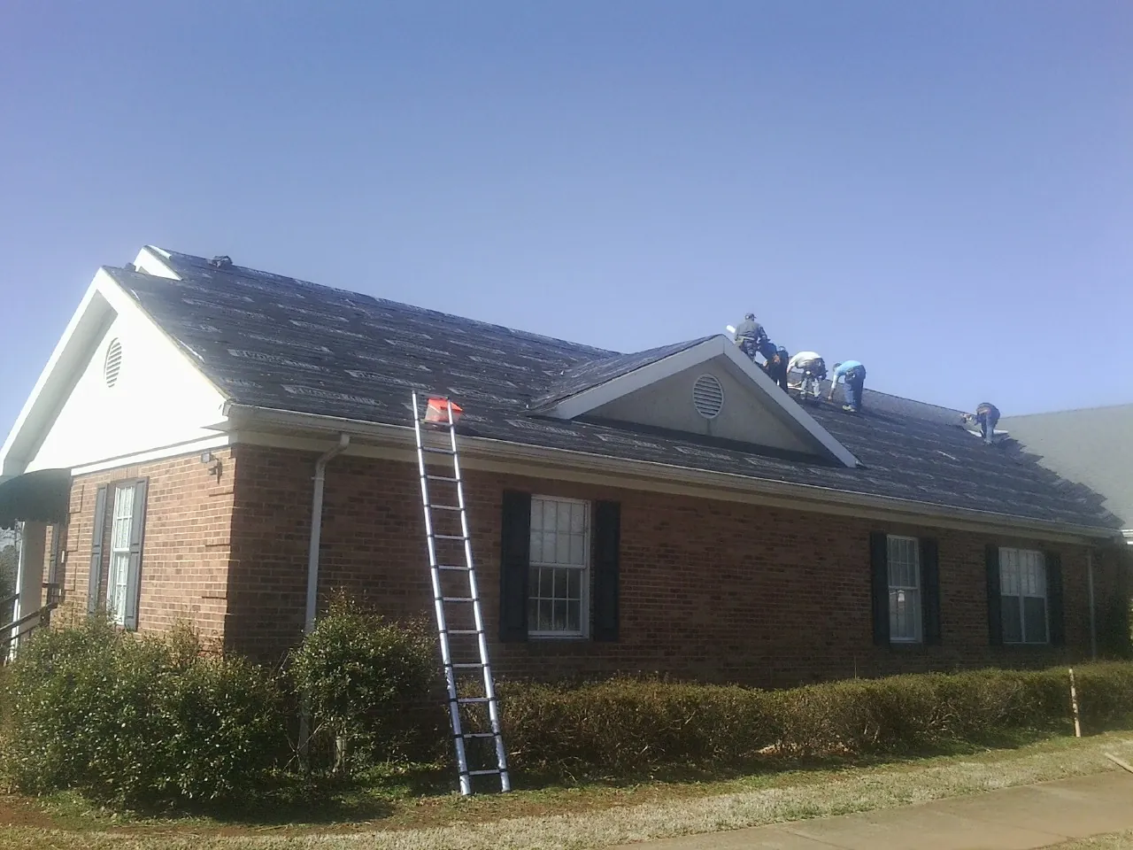 Crew tearing shingles off a brick ranch roof with a ladder set — Emergency Roof Repair work by our crew in Conehatta