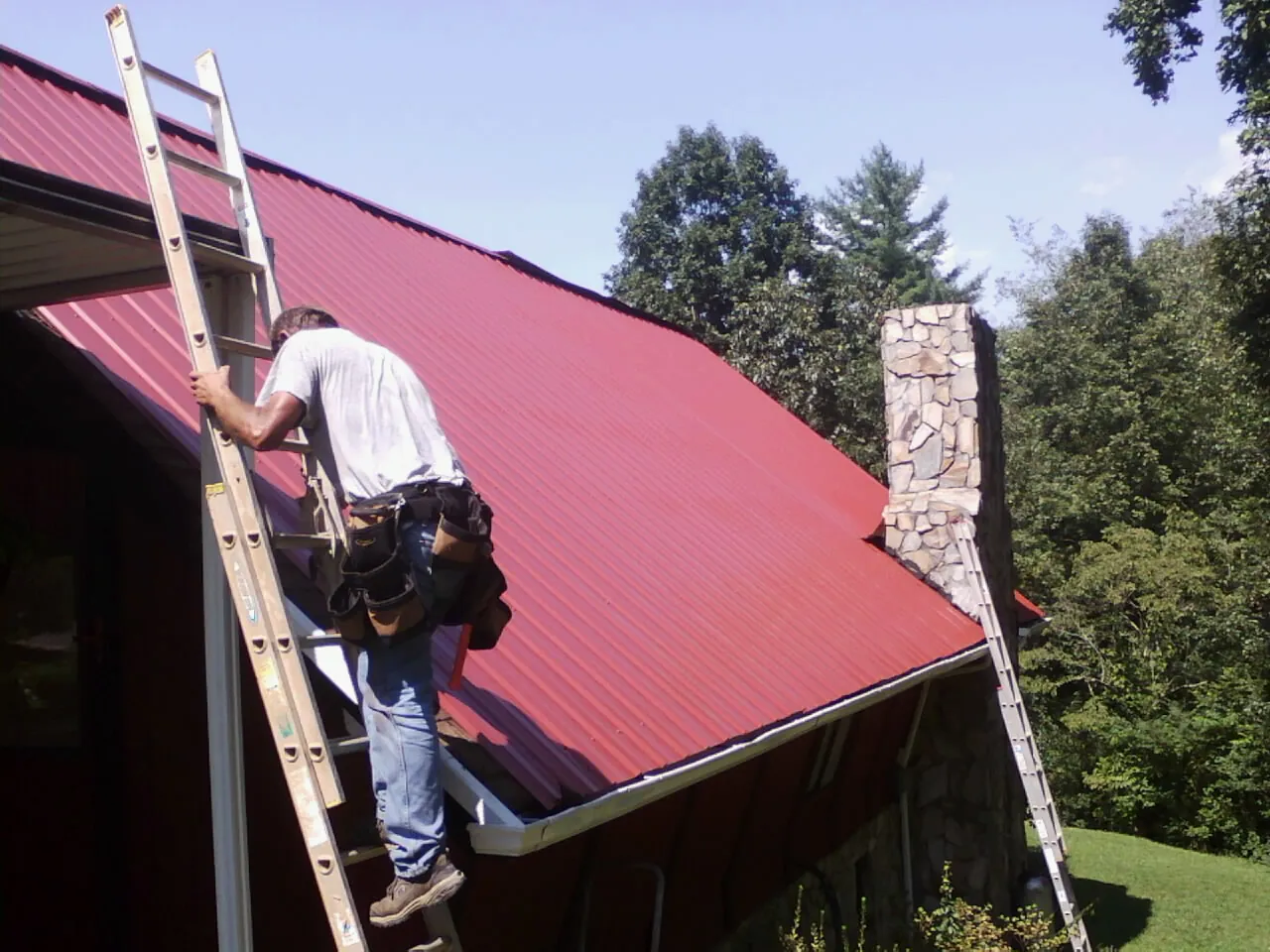 Roofer on a ladder beside a red metal roof and stone chimney — Soffit & Fascia Repair by Roofing Brothers in Conehatta