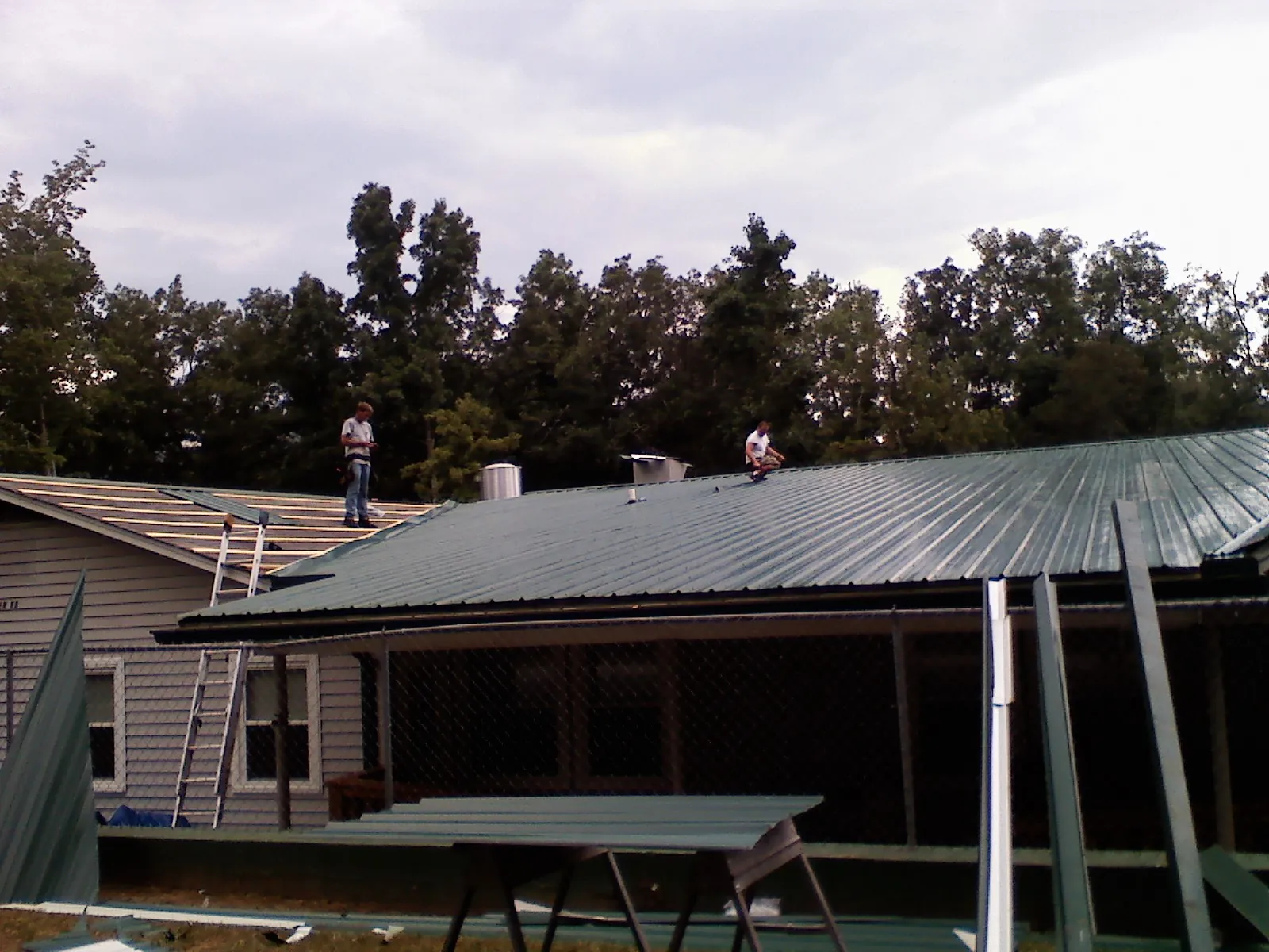 Two roofers installing green metal panels above a covered porch in Conehatta